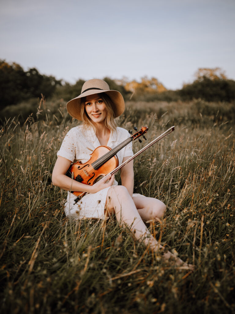 Olivia Barrett sits with her fiddle in the long grass.